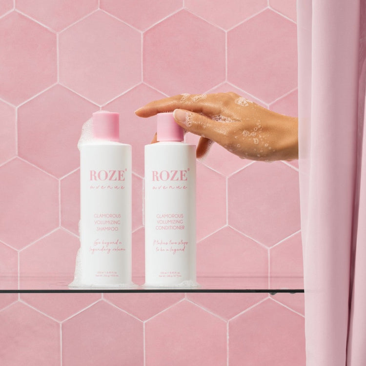 szampon i odżywyka Roze Avenue - glamorous volumizing shampoo i conditioner ustawione obok siebie na szklanej półce, ręka kobiety spod prysznica sięga po odżywkę