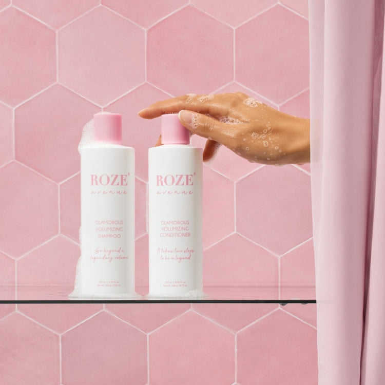 szampon i odżywyka Roze Avenue - glamorous volumizing shampoo i conditioner ustawione obok siebie na szklanej półce, ręka kobiety spod prysznica sięga po odżywkę