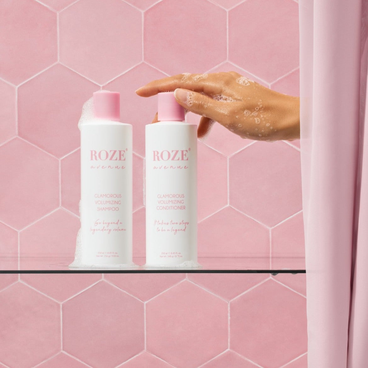 szampon i odżywyka Roze Avenue - glamorous volumizing shampoo i conditioner ustawione obok siebie na szklanej półce, ręka kobiety spod prysznica sięga po odżywkę